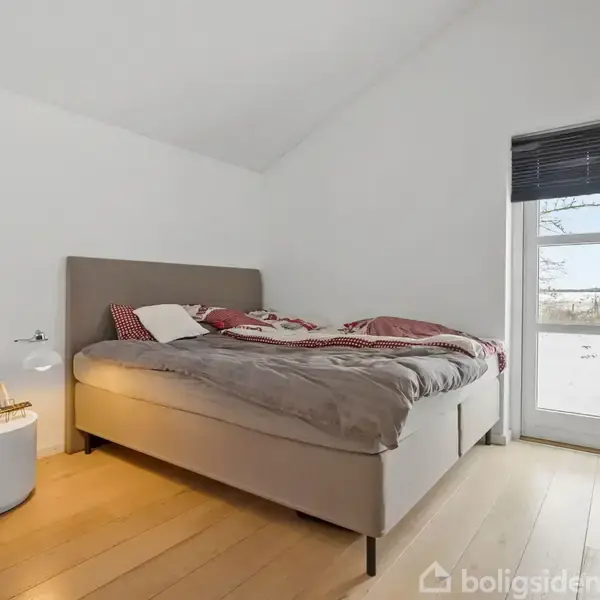 Seng med beige gavl står i et lyst soveværelse. Sengens dyne er uredt med rødt mønster. Ved siden af står et rundt natbord med lampe. Vindue viser snedækket landskab.