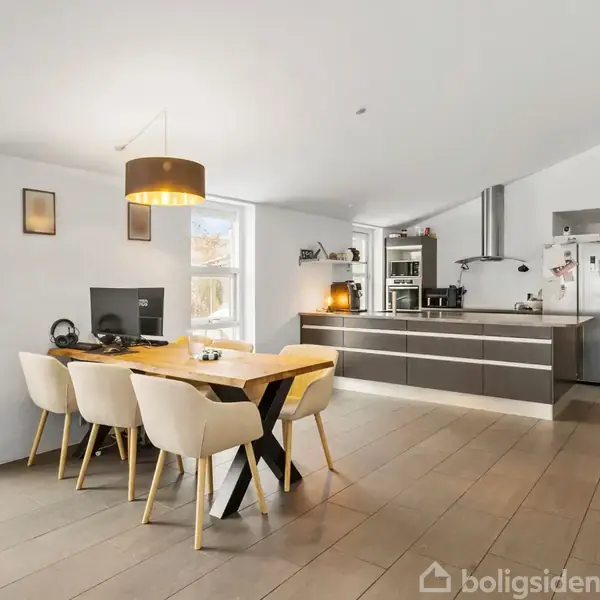 Dør står åben i en smal gang med gråt køkkenelement og vaskemaskine. Vask og opbevaring til højre, lysdøre fører til næste rum. Gulv er flisebelagt, vægge er hvide.