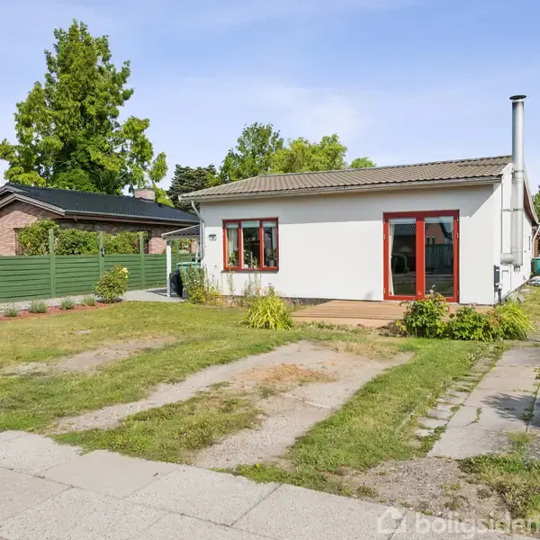 Huset er enkelt og moderne med fladt tag og store vinduer. Omkringliggende grønne planter og en uplejet forhave med fliser giver et afslappet, naturligt udendørsmiljø.