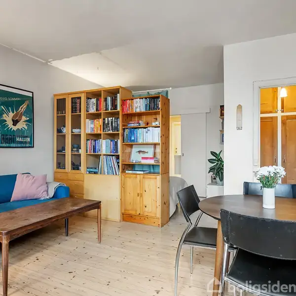 Reol med bøger står i en stue, mens et rundt bord med stole befinder sig i forgrunden. En sofa og kunst hænger på væggen; teksten på plakaten er "Bjerg Maleren".