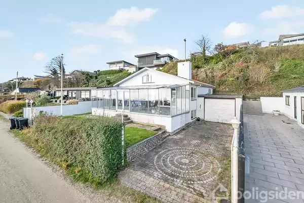 Hus med hvid facade og glaseret veranda, står på en brolagt gårdsplads omgivet af buske. Bakkede omgivelser med andre huse i baggrunden.