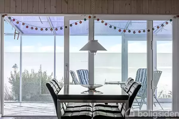 Et bord med sorte stole står indendørs foran en glasdør. Udenfor er der en terrasse med farverige lyskæder og havemøbler, med udsigt over en klar himmel.