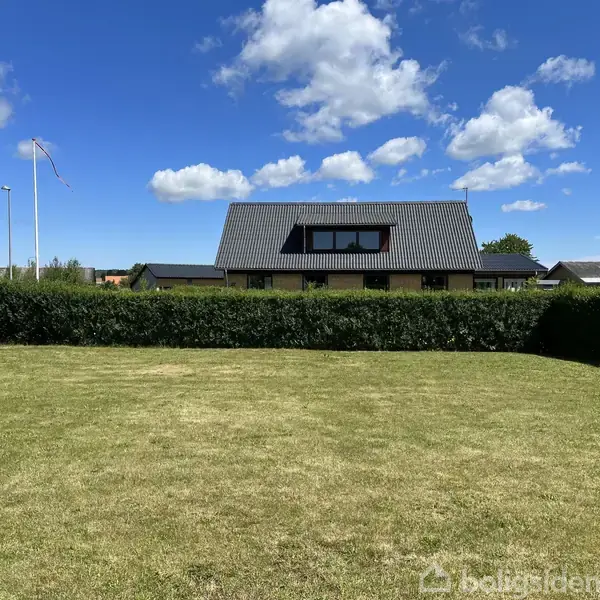 Et moderne hus med grå tag bevogtes af en tæt hæk i en velplejet have, under en blå himmel med hvide skyer. En flagstang står til venstre.