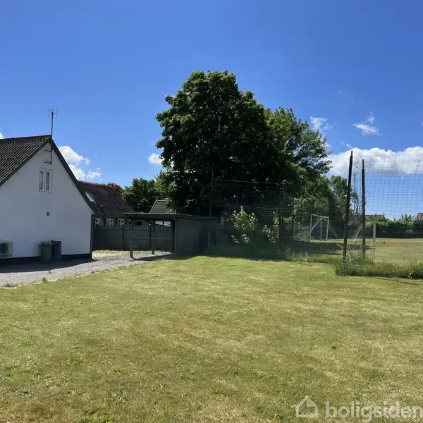 Et hvidt hus står stille ved siden af en velplejet græsplæne, omgivet af en fodboldbane med mål til højre og grønne træer i baggrunden under en blå himmel.