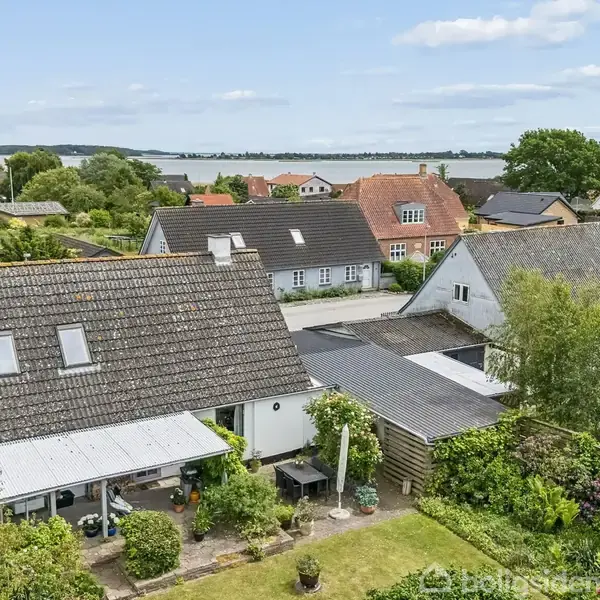 Et rækkehus med mørkt tag står stille i en villahave omgivet af frodig vegetation og andre huse. I baggrunden ses en sø eller fjord under en klar, blå himmel.