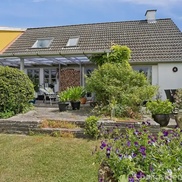Hus med skråt tag og flere vinduer, omgivet af en frodig have med blomster og buske. En terrasse med møbler ses foran huset under en pergola med brænde stablet til højre.