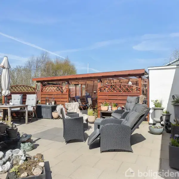 Havemøbler står på en terrasse omgivet af blomsterkrukker og stakit. Et bord med parasol og stole ses i baggrunden, under en blå himmel med nogle få skyer.