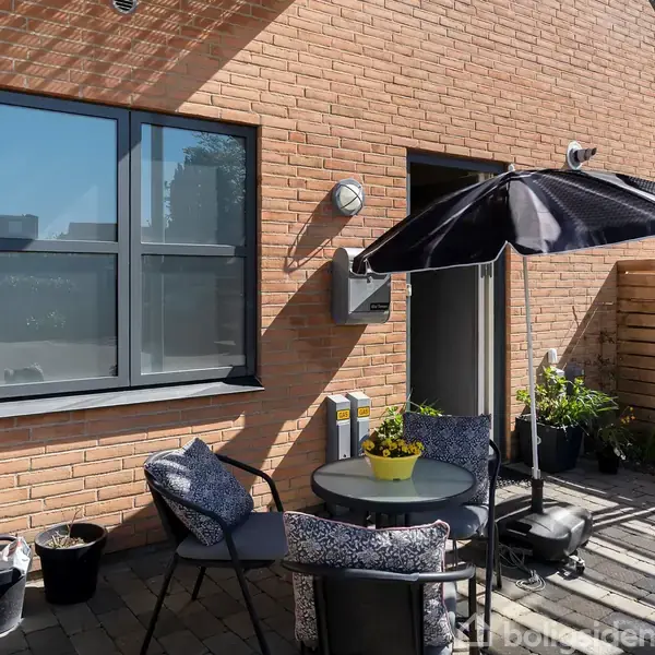 Rundt havebord med fire stole, der står under en sort parasol. Omgivet af røde murstensvægge, krukker med planter og en træskillevæg på en terrasse.