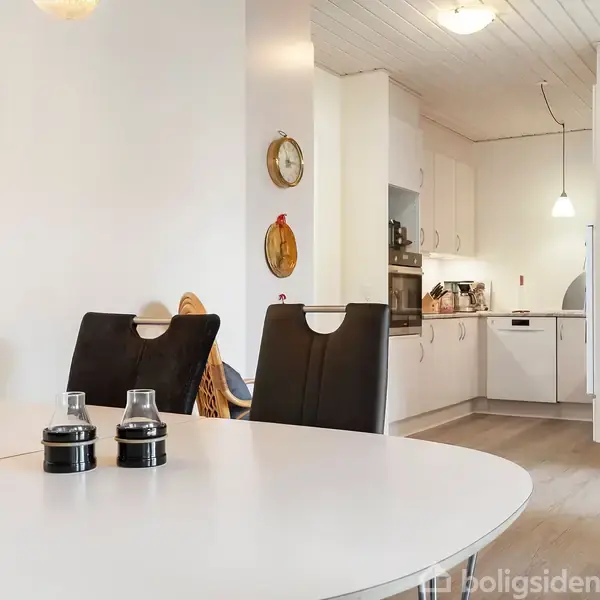 Rundt spisebord med krydderikrukker står i en moderne spisestue. To sorte stole ved bordet. I baggrunden ses et lyst køkken med ophængte lamper og en åben skab med glaslåger.