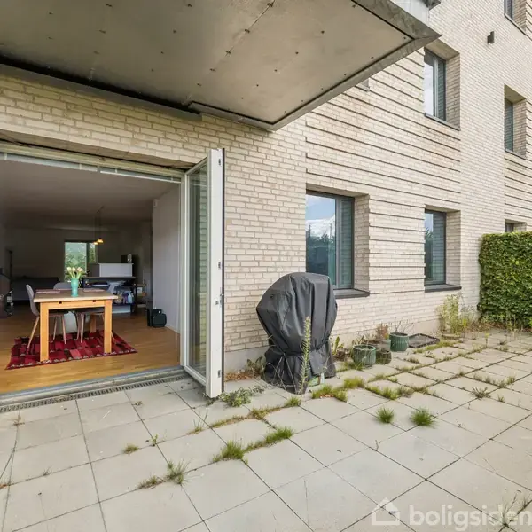 En terrasse med fliser og græs mellem dem foran en moderne bygning. Dobbeltdøre står åbne til en stue med spisebord. En grill er dækket til ved siden af.