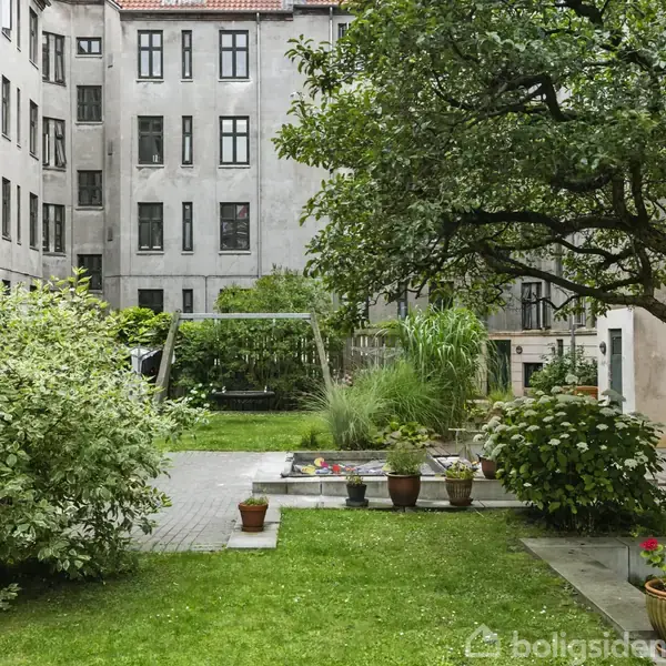En stue med en beige sofa og glasbord, omgivet af bøger på en hylde. Væggene er lyseblå med et billede hængende. Der er en gulvlampe og grønne planter.