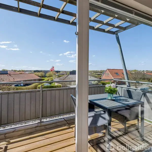 Bord med stol står på en balkon med træterrasse. Udsigt over villakvarter med røde hustage og et dansk flag i baggrunden under en blå himmel med lette skyer.