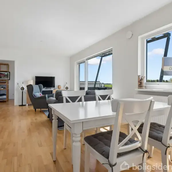Spisebord med fire stole står ved vindue i lys stue. Sofa og tv er placeret i baggrunden, omkranset af træpaneler og kunstværker på væggene.