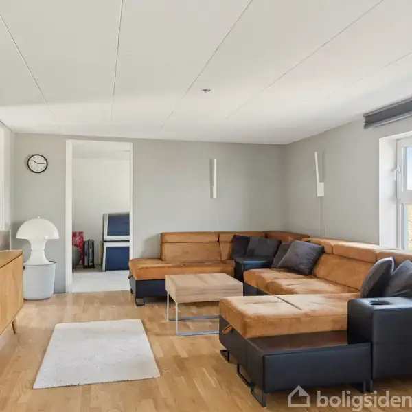 En stor, L-formet brun sofa med mørke puder står i en lys stue med trægulv. Et hvidt tæppe og et træbord er placeret foran sofaen. En stor vindue slipper naturligt lys ind.