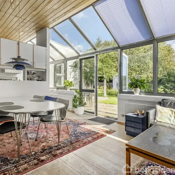 Et lyst køkken-alrum med et rundt spisebord og sorte stole står på et orientalsk tæppe. Rummet har store vinduer mod haven, og planter skaber en hyggelig atmosfære.