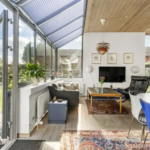 En stue med store vinduer og døre åbne mod en have. En sofa og bord er placeret på et tæppe nær en TV-skærm på væggen. Planter og moderne møbler kompletterer rummet.