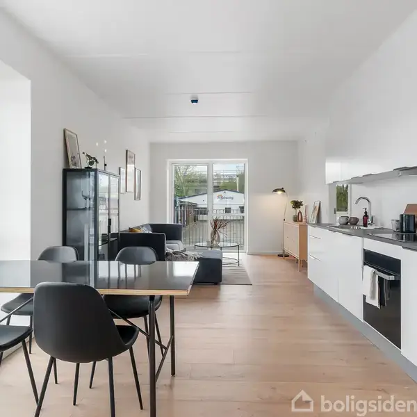 Spisebord med sorte stole står i moderne, lys stue-køkken. Køkkenelementer i hvidt langs højre væg. Store vinduer giver udsigt til terrasse. Skab og sofa ses i baggrunden.
