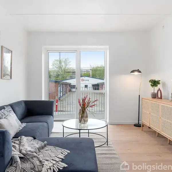 Sofa er placeret ved siden af et glasbord med en vase, i en stue med lysindfald fra store vinduer, dekoreret med kunst på væggene og en trækommode ved siden.