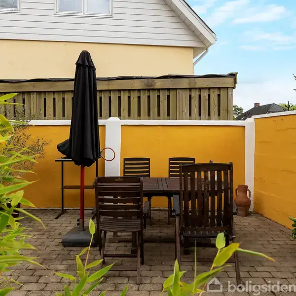 Terrassemøbler står på en flisebelagt gårdhave. En parasol er foldet sammen ved siden af bordet. Omringet af gule vægge og planter foran et hus med gul facade og skråt tag.