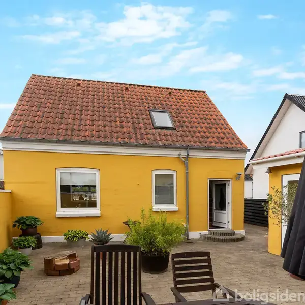 Gulmalet hus med rødt tegltag, omgivet af en brostensbelagt gårdhave med planter. En parasol og liggestole står ved siden af huset under en blå himmel.