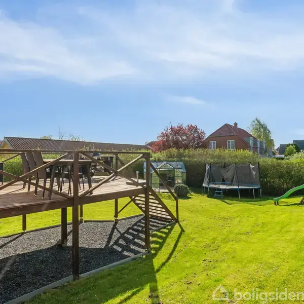 En træterrasse med stole og bord står ved et hus. I haven er der en trampolin og en rutsjebane. Omkring dem ses en velplejet græsplæne og buskads.