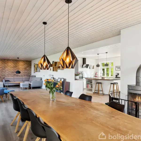 Et stort spisebord med blomster i midten står i en moderne spisestue. Flot belysning hænger over bordet. I baggrunden ses en stue med sofaer og en væg med mursten. En brændeovn står til højre.