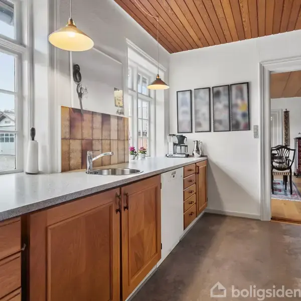 Et køkken viser en vask og bordplade i lyst træ med skabe. En kaffemaskine står ved siden af dekorative billeder på væggen. Ved siden ses en stue med træmøbler.