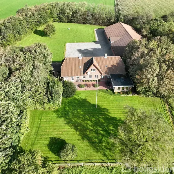 En stor villa med rødt tag ligger midt i en frodig, grøn have omgivet af træer. Der er en tilknyttet udbygning bagved, omgivet af en velplejet græsplæne.