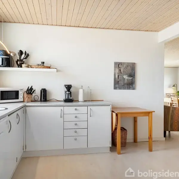 Et lyst køkken med hvide skabe og stålgreb, står ved siden af en træbord. Kaffemaskiner og køkkenredskaber er på bordet. Et skilt med "CAFE" hænger på væggen.