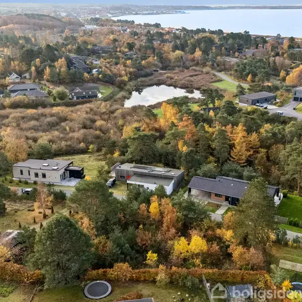 Boliger ligger blandt træer i et efterårslandskab med en sø i baggrunden, omgivet af skovklædte områder, tæt på en fjord.