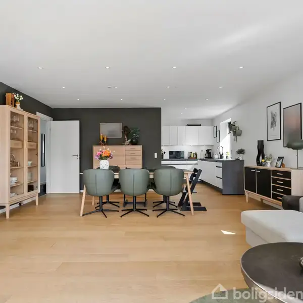Stue med spisebord og seks stole i midten, omgivet af moderne møbler og en åben forbindelse til køkkenet. Rummet er lyst og enkelt indrettet med billeder på væggene.