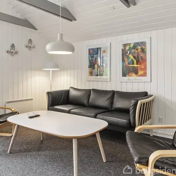 Sort lædermøbelarrangement med sofa og to stole omkring et hvidt træbord i en stue. Lysegrå panelvægge med kunstplakater og moderne belysning skaber en hyggelig atmosfære.