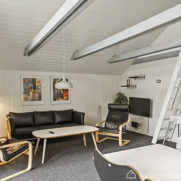 Stue med sort lædersofa og stole, kaffebord foran, lampeskærm hængende over bordet, stige til loftet i baggrunden, hvide vægge, og billeder på væggen i et moderne rum.
