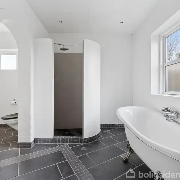 Et fritstående badekar står i et lyst badeværelse med sort flisegulv; en åben bruseniche er til venstre, og et vindue mod haven giver naturligt lys.