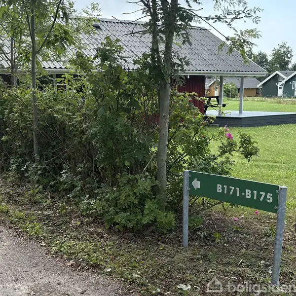 Et grønt skilt viser vej til B171-B175 blandt buske, ved siden af en terrasse og en rød bygning i en have med græs.