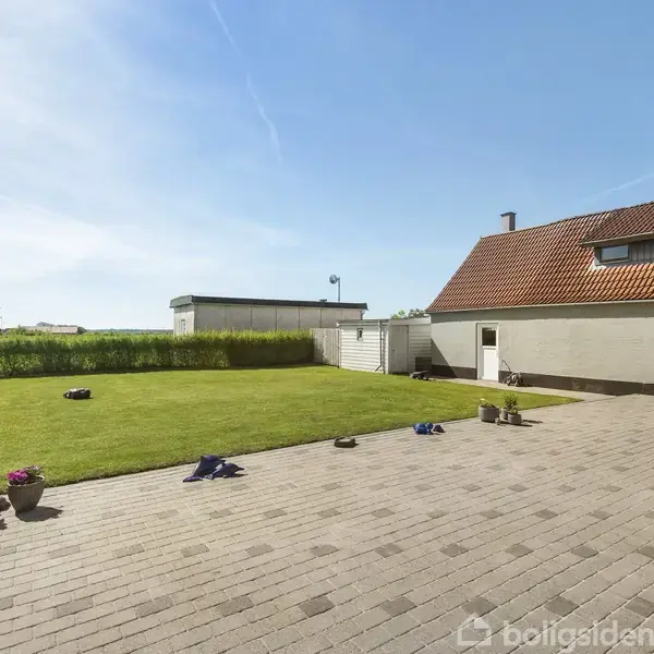 Græsplæne omgivet af en flisebelagt terrasse med blomsterkrukker, ved siden af et hus med rødt tag og store vinduer, i et åbent, solrigt landskab.