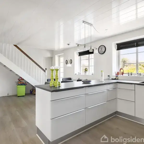 Køkken med hvide skabe og sort bordplade, beliggende i et lyst rum med trægulve. Store vinduer giver naturligt lys. Trappe i baggrunden fører opad. To vægure hænger over vasken.