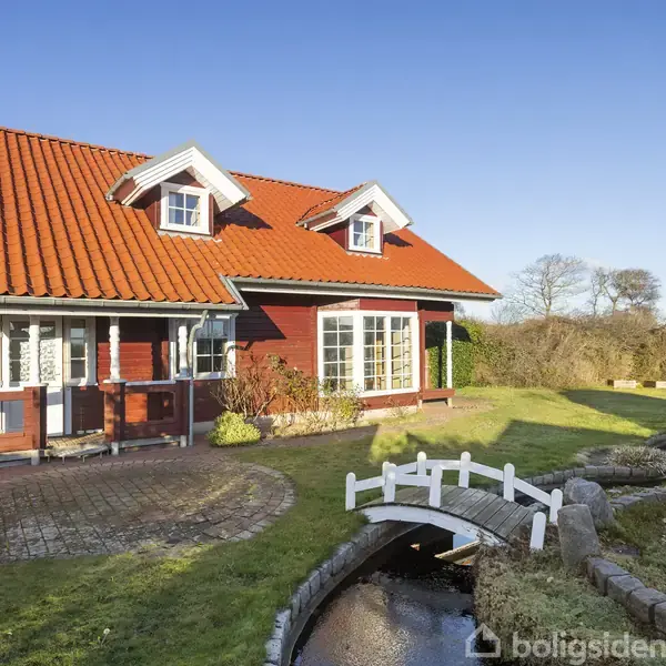 Et rødt hus med hvide vinduer og rødt tag står stille i en velplejet have. Foran huset krydser en lille hvid bro et smalt vandløb.
