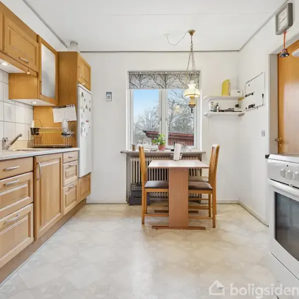 Køkken med træskabe og hvide fliser, hvor en elkedel står på bordet. Et spisebord med stole er placeret ved vinduet. Rummet er lyst og rummeligt.