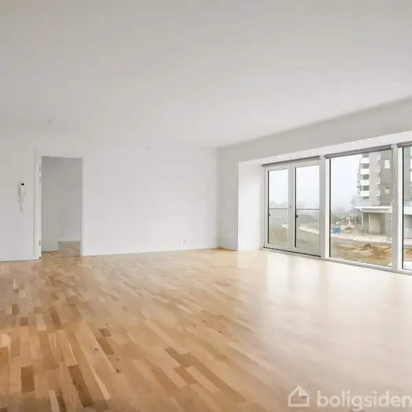 Lyst rum med trægulv, store vinduer mod moderne bygninger udenfor. Rummet er tomt, hvilket giver en fornemmelse af rummelighed.