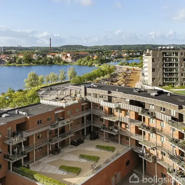 Rød murstensbygning med altan foroven, glasrækværk og flere vinduer. I forgrunden står et havebord med stole på en terrasse af fliser og træ.