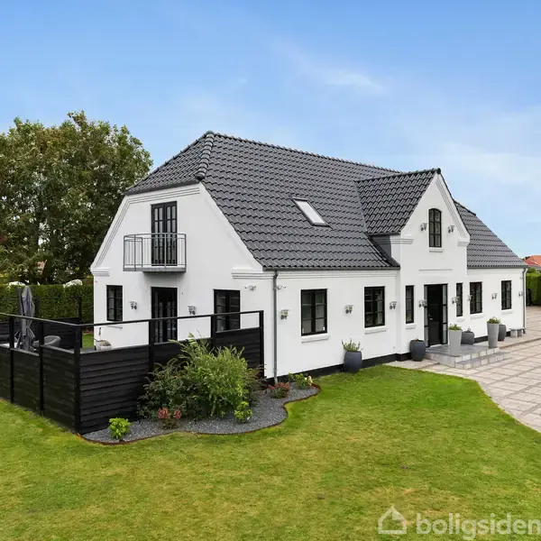 Hvidt hus med sort tag og mange vinduer, omkranset af en terrasse med havemøbler. Huset står i en grøn have med en sort indhegnet område til venstre og flisebelagt areal til højre.