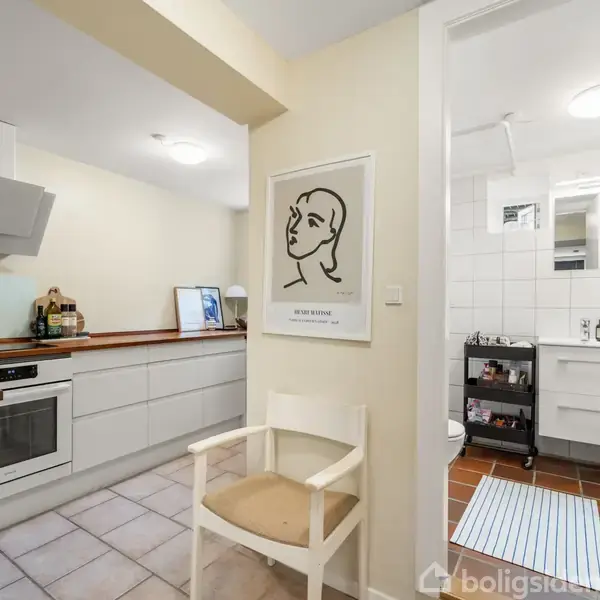 En moderne køkken med hvide skabe og integreret ovn. En stol står ved en væg med en minimalistisk kunstplakat. Et tilstødende badeværelse ses gennem en åben dør.