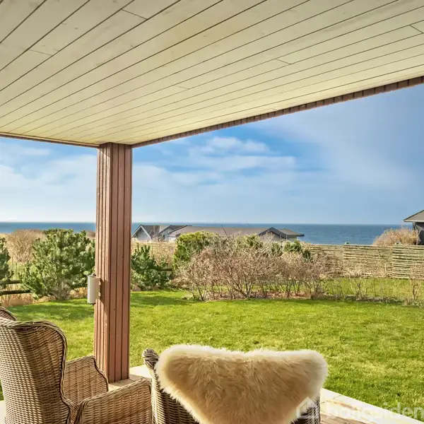 En terrasse med flettede stole, delvis overdækket, giver udsigt til en grøn have og havet i baggrunden. Små buske og hække fungerer som skel til nabohuse.