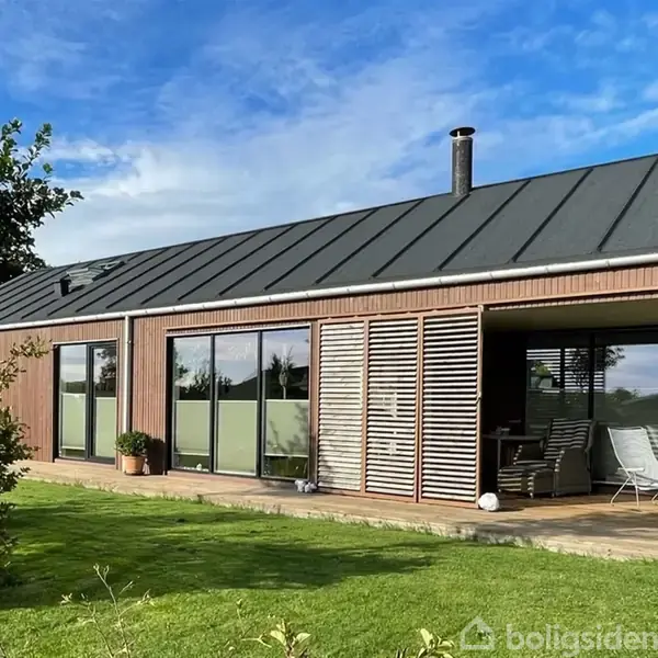 Et moderne, træbeklædt hus med store vinduespartier og en terrasse. Det står i en velplejet have med græsplæne og træer. Himlen er delvist skyet med blå nuancer.