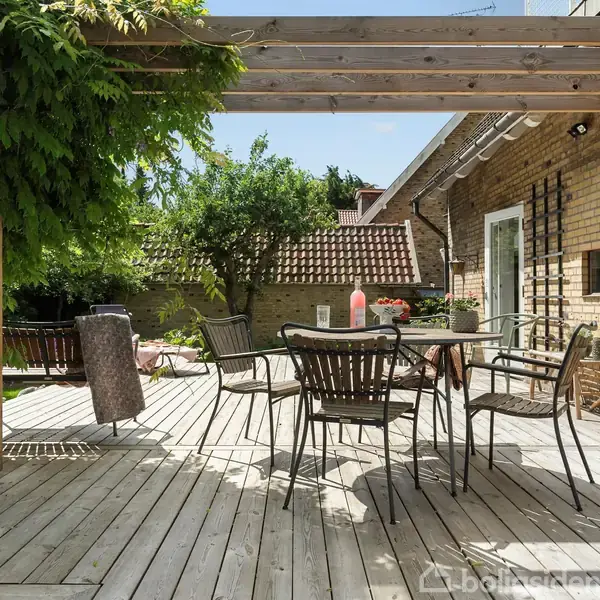 Terrassemøbler er arrangeret på en træterrasse under en pergola med grønne planter, omgivet af en murstensbygning og en have med træer. Sollys skaber en afslappet atmosfære.
