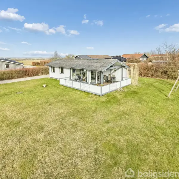 Et sommerhus står stille på en stor græsplæne med en terrasse og en gynge ved siden af. Omgivet af buske og træer under en blå himmel med skyer.