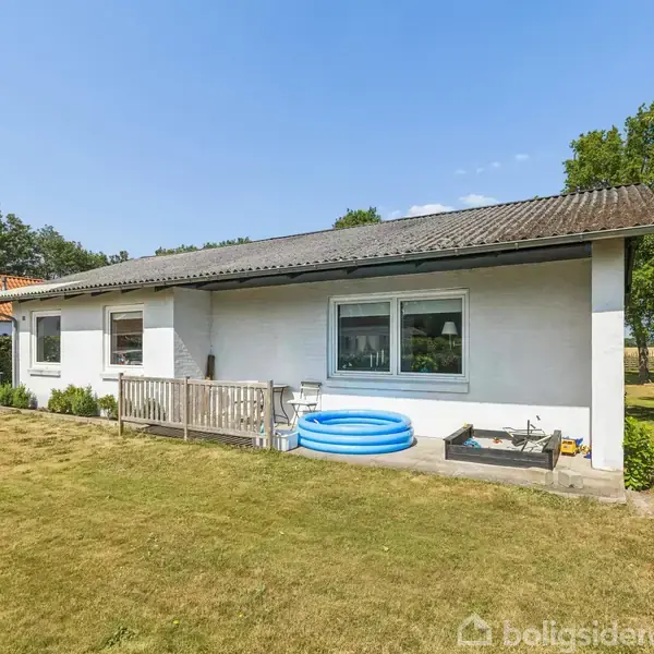 Et hvidt enfamiliehus med fladt tag står i en have med græsplæne. En blå oppustelig pool ses foran huset, omgivet af buske og træer under en klar blå himmel.