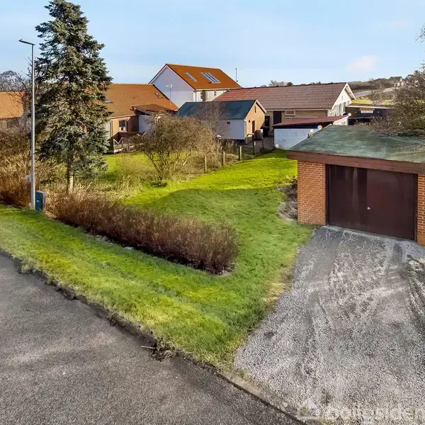 Garagebygning med brun port står i en have med græs og buske, beliggende ved en asfalteret vej. I baggrunden ses flere huse med rødt tag i et kvarter.