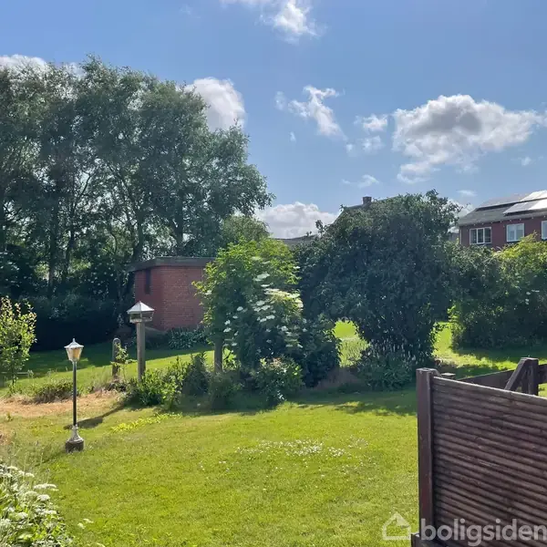 Græsplæne med røde parasoller og lamper i en have. Et rødt murstenshus ses i baggrunden omgivet af buske og træer under en blå himmel med skyer.